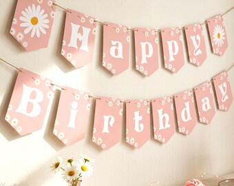 May include: A pink birthday banner featuring white daisy flower patterns and large white letters spelling out Happy Birthday. The banner is composed of two rows of pennant-shaped cards strung together on a thin brown twine cord.
