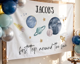 May include: A white rectangular banner featuring the text "JACOB'S first trip around the sun" in black lettering. The design includes watercolor illustrations of planets, a rocket ship, and gold glitter stars, framed by a wooden stand with balloons.