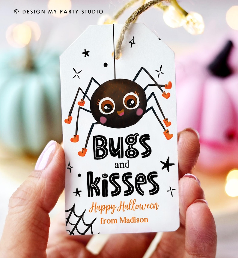 May include: White Halloween gift tag with a cartoon spider illustration. The tag reads "Bugs and Kisses" and "Happy Halloween from Madison". The spider is brown with orange accents. The tag is held by a person.