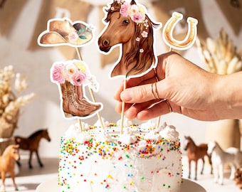 May include: A set of four decorative cake toppers featuring a brown horse head with a floral crown, a pair of brown cowboy boots filled with pink and white flowers, a brown cowboy hat with flowers, and a gold horseshoe, all attached to sticks.