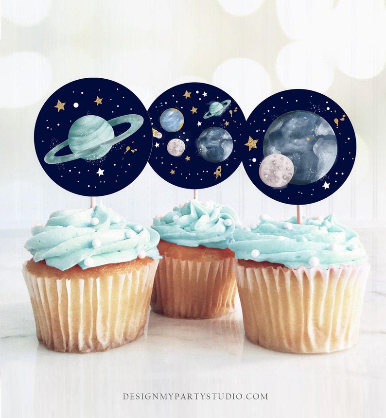 May include: Three blue and white cupcake toppers with a space theme. The toppers feature planets, stars, and a rocket ship on a dark blue background.