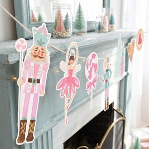 Op de afbeelding: Een decoratieve slinger met papieren uitsparingen van een notenkraker, een ballerina, een lolly, een muis, een dennenboom en een hart, allemaal geregen aan een lichtgekleurd touw. De illustraties bevatten zachte roze, blauwgroene en bruine tinten.