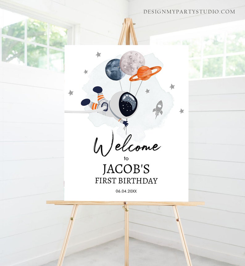May include: Welcome to Jacob's First Birthday sign with a blue and white astronaut holding onto balloons shaped like planets. The sign has a watercolor background with stars and a rocket.