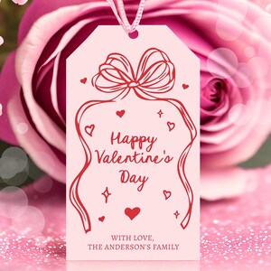 Puede incluir: Etiqueta de regalo rosa con un lazo rojo y un dise&ntilde;o de lazo. El texto dice "Feliz d&iacute;a de San Valent&iacute;n" con un coraz&oacute;n rojo y las palabras "Con amor, La familia Anderson".