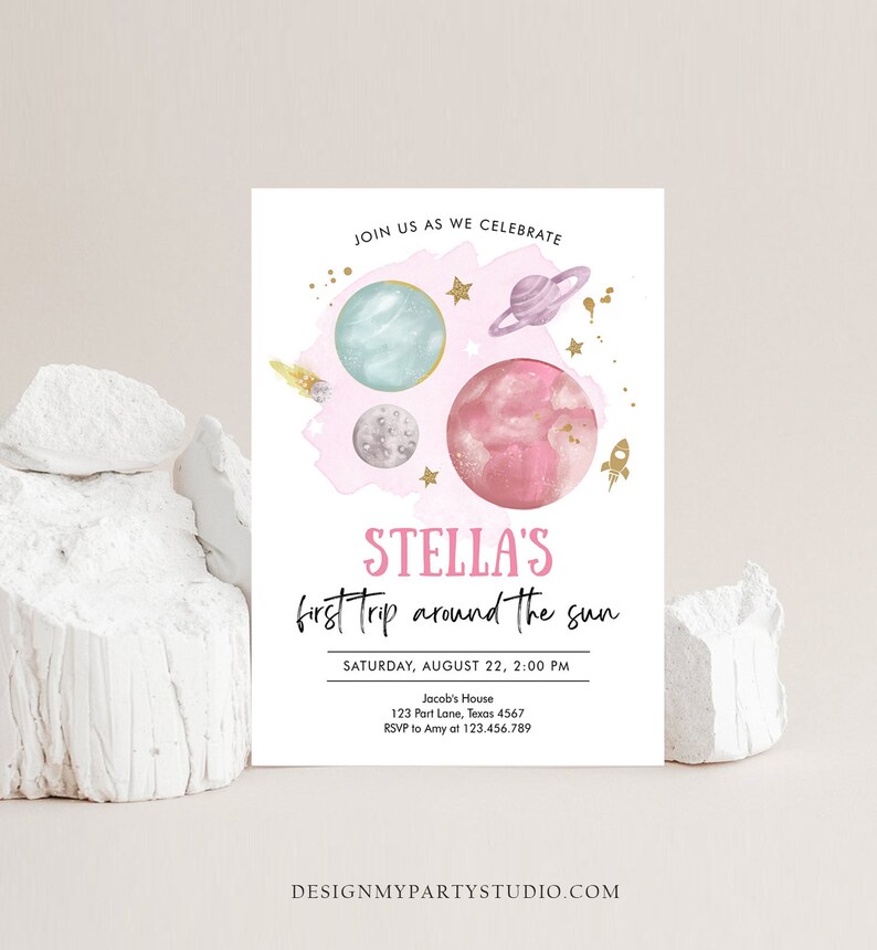 May include: A pink and blue space-themed invitation for a child's first birthday party. The invitation features planets, stars, and a rocket ship. The text on the invitation reads "Stella's first trip around the sun" and includes the date, time, and location of the party.
