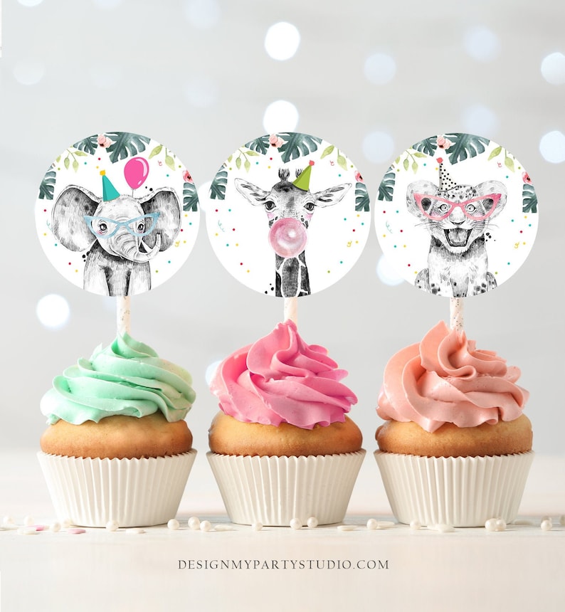 May include: Three cupcakes decorated with animal-themed toppers. The toppers are round and feature an elephant wearing glasses, a giraffe blowing a bubble, and a lion wearing sunglasses. The cupcakes are frosted in green, pink, and peach.