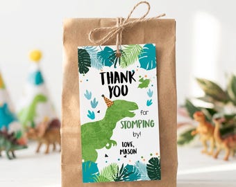May include: A brown paper gift bag with a rectangular tag tied by twine. The tag features a green dinosaur wearing a party hat, surrounded by tropical leaves and the text: THANK YOU for STOMPING by! LOVE, MASON. The design includes blue and green accents.
