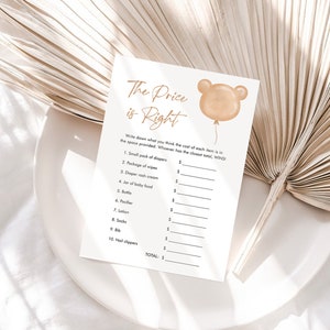 May include: A baby shower game card titled "The Price is Right" with a bear-shaped balloon graphic. The card lists baby items like diapers and lotion, with spaces to write prices. The card is on a white plate with a neutral background.