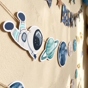Op de afbeelding: Een ruimteslinger met uitgesneden figuren van een astronaut in een wit en blauw pak, een turquoise planeet met ringen en diverse donkerblauwe en grijze hemellichamen, bevestigd aan een dun bruin touw voor decoratieve ophanging.