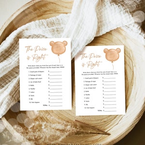 May include: Two baby shower game cards with the text "The Price is Right" and a bear balloon graphic. The cards are on a wooden tray with a white cloth. The game includes a list of baby items to price.