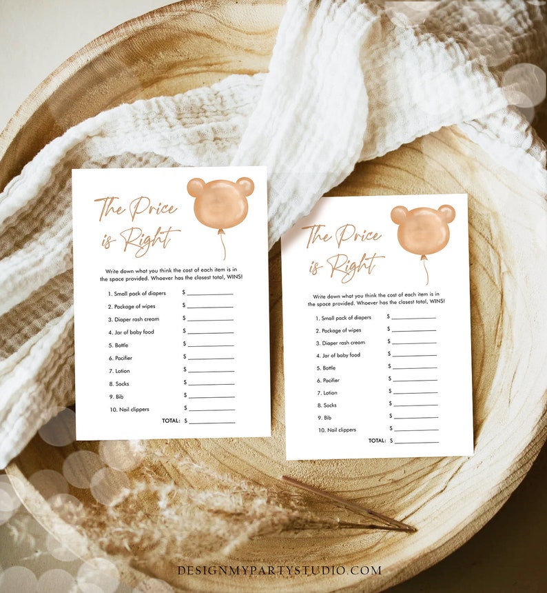 May include: Two baby shower game cards with the text "The Price is Right" and a bear balloon graphic. The cards are on a wooden tray with a white cloth. The game includes a list of baby items to price.