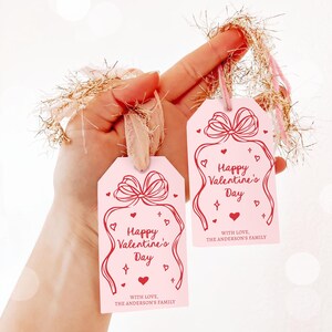 Puede incluir: Dos etiquetas de regalo rosas con texto rojo que dice "Feliz d&iacute;a de San Valent&iacute;n" y "Con amor, la familia Anderson". Cada etiqueta tiene un contorno rojo de un lazo con corazones y estrellas.