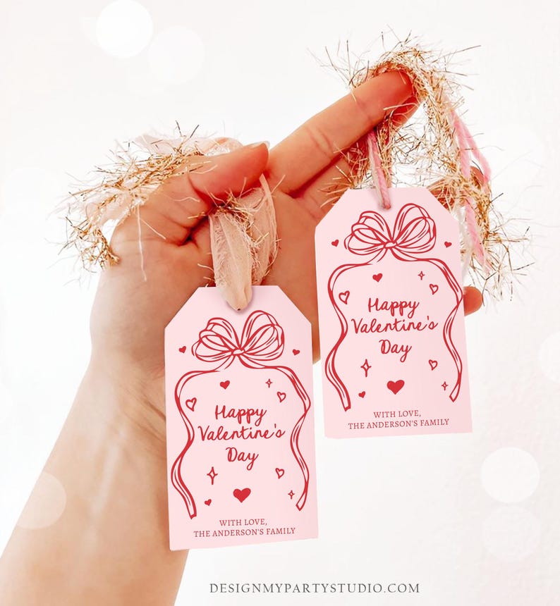 Puede incluir: Dos etiquetas de regalo rosas con texto rojo que dice "Feliz d&iacute;a de San Valent&iacute;n" y "Con amor, la familia Anderson". Cada etiqueta tiene un contorno rojo de un lazo con corazones y estrellas.