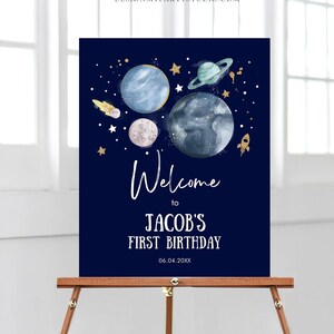 May include: A blue and gold welcome sign with a space theme. It says "Welcome to Jacob's First Birthday 06.04.20XX". The sign features planets, stars, and a rocket.