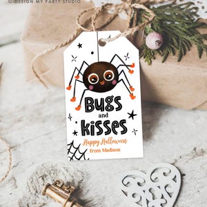 Pu&ograve; includere: Un'etichetta regalo rettangolare bianca con l'illustrazione di un ragno marrone con grandi occhi e accenti arancioni. Il testo recita Bugs and kisses, Happy Halloween e from Madison. Include grafiche di ragnatele e stelle nere.