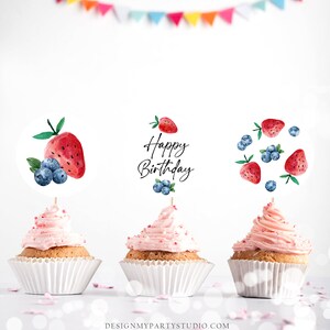 May include: Three pink frosted cupcakes with white cupcake liners. Each cupcake has a different watercolor fruit themed topper. One topper says "Happy Birthday" with blueberries and strawberries. Another topper has a strawberry and blueberries. The last topper has a watermelon and blueberries.
