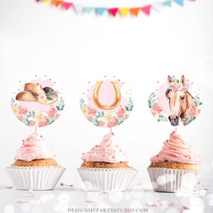 May include: Three pink cupcakes with white frosting and sprinkles. Each cupcake has a round topper with a pink floral design. The toppers feature a cowboy hat, a horseshoe, and a horse's head.