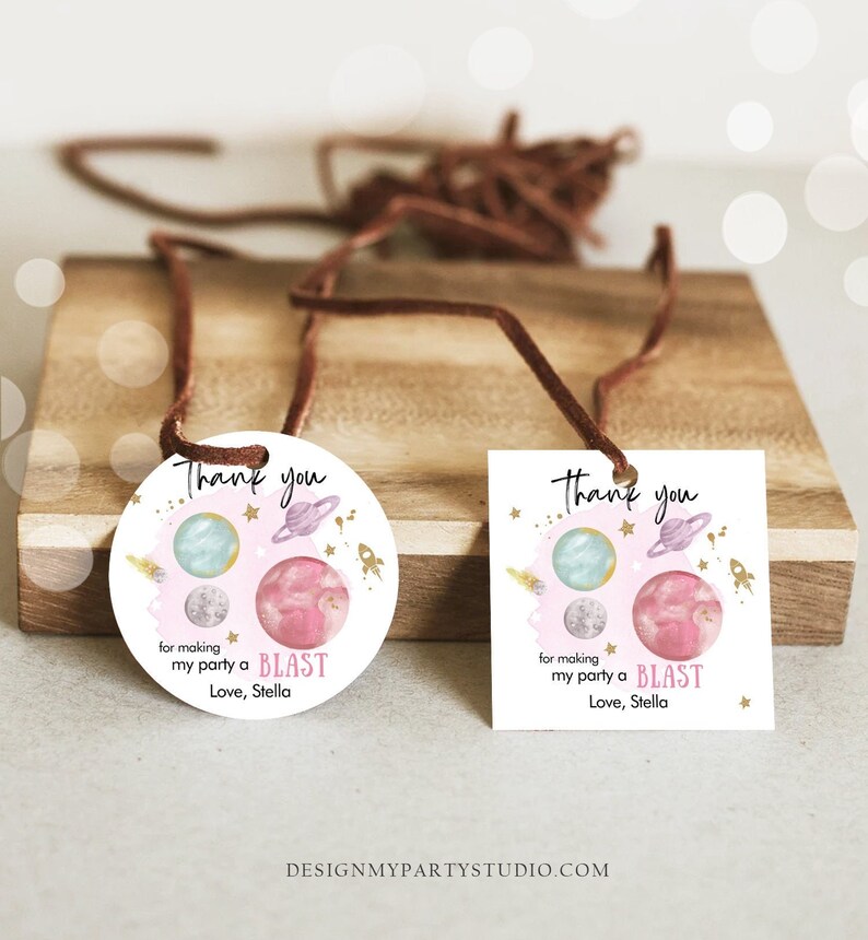 May include: Two round and square thank you tags with a space theme. The tags feature a pink and blue watercolor galaxy with planets, stars, and a rocket ship. The text on the tags reads "Thank you for making my party a BLAST Love, Stella".