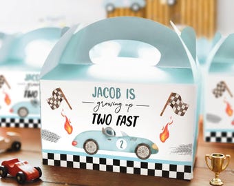Op de afbeelding: Een lichtblauw kartonnen doosje met handvat in racewagenthema. De doos bevat de tekst "JACOB IS growing up TWO FAST" met illustraties van een lichtblauwe racewagen met het nummer twee, geblokte vlaggen, vlammen en bandensporen op de onderkant.