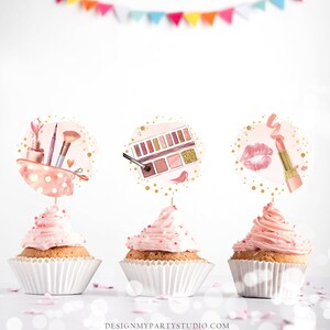Puede incluir: Tres cupcakes rosas con glaseado blanco y chispas doradas. Cada cupcake tiene una tapa redonda de papel blanco con un dise&ntilde;o diferente de tem&aacute;tica de maquillaje: una bolsa de maquillaje con pinceles y esmalte de u&ntilde;as, una paleta de sombras de ojos con una brocha y un l&aacute;piz labial con una marca de beso.