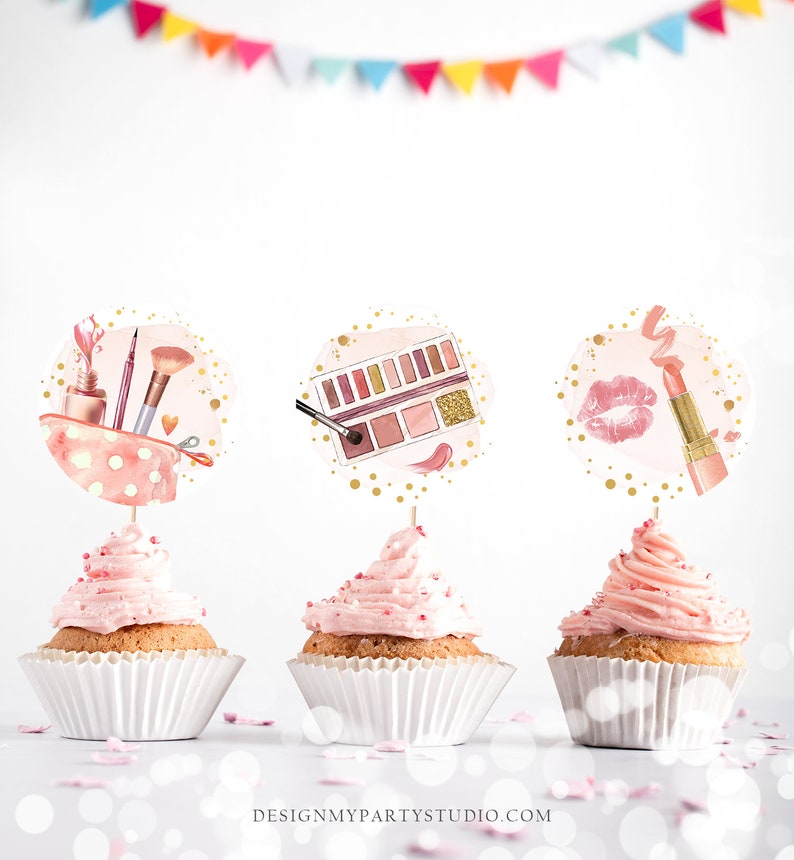 Puede incluir: Tres cupcakes rosas con glaseado blanco y chispas doradas. Cada cupcake tiene una tapa redonda de papel blanco con un dise&ntilde;o diferente de tem&aacute;tica de maquillaje: una bolsa de maquillaje con pinceles y esmalte de u&ntilde;as, una paleta de sombras de ojos con una brocha y un l&aacute;piz labial con una marca de beso.