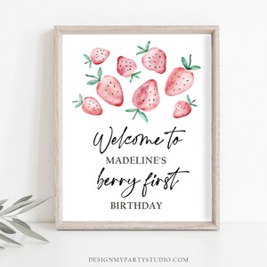 May include: A white sign with a wood frame and a watercolor illustration of strawberries. The sign says "Welcome to Madeline's berry first birthday".