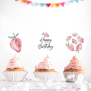 May include: Three pink cupcakes with white frosting and red sprinkles. Each cupcake has a different strawberry-themed topper. One topper says "Happy Birthday" in black script, another has a single red strawberry, and the last topper has a cluster of red strawberries.