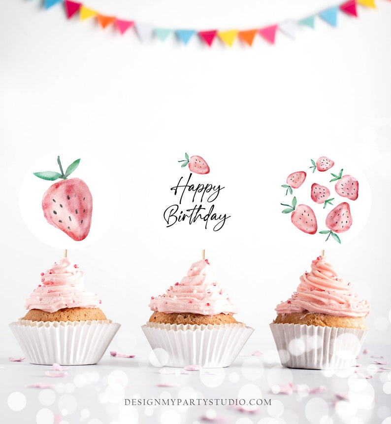 May include: Three pink cupcakes with white frosting and red sprinkles. Each cupcake has a different strawberry-themed topper. One topper says "Happy Birthday" in black script, another has a single red strawberry, and the last topper has a cluster of red strawberries.