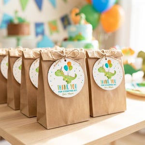 May include: A row of brown paper gift bags, each featuring a round white tag tied with twine. The tag displays a green cartoon dinosaur wearing a party hat, holding three colorful balloons, with the text Thank You For Stomping By! Love, Mason.