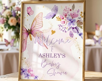 May include: A rectangular gold-framed sign featuring watercolour illustrations of pink and purple butterflies and floral bouquets. The text reads Welcome to ASHLEY'S Baby Shower in a mix of script and block lettering against a soft pink background.