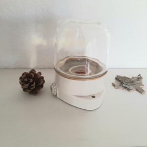 Könnte beinhalten: Weißer Keramik-Luftbefeuchter mit einem klaren Glasdeckel. Der Luftbefeuchter hat einen goldenen Rand oben und unten.