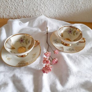 Könnte beinhalten: Zwei Vintage-Tassen und Untertassen mit Goldrand und floralen Designs. Die Tassen und Untertassen sind cremefarben mit zarten rosa und gelben Blumenmustern. Ein Zweig rosa Blüten liegt zwischen den Tassen.