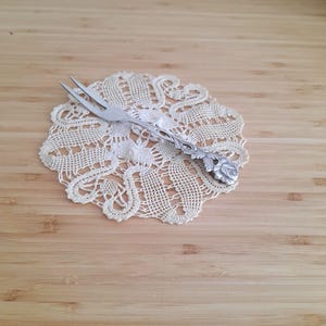 Peut inclure: Une fourchette à gâteau en argent avec un motif de rose sur le manche. La fourchette repose sur un napperon en crochet blanc.