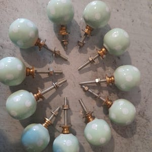 Puede incluir: Una colección de diez pomos de cerámica verde claro con lunares blancos. Cada pomo tiene un tornillo y una tuerca de metal para la instalación. Los pomos están dispuestos en un patrón circular sobre una superficie gris.
