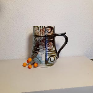 Könnte beinhalten: Ein Keramikbecher mit schwarzem Henkel und einzigartigem Design. Der Becher weist eine Mischung aus Farben auf, darunter Braun, Blau und Weiß, mit abstrakten Mustern. Ein kleiner Strauß orangefarbener Beeren liegt in der Nähe des Becherfußes.