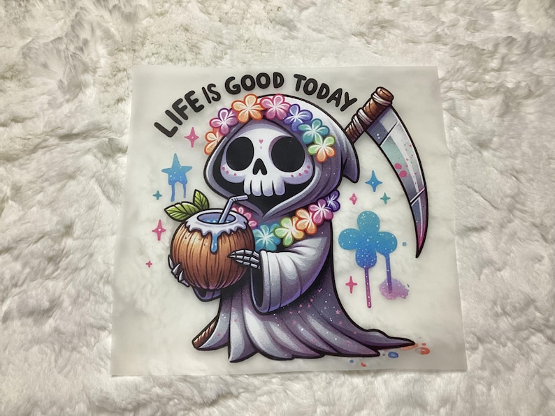 Puede incluir: Una imagen de dibujos animados de la Parca con una corona de flores y sosteniendo un coco con una pajita. El texto "LIFE IS GOOD TODAY" est&aacute; por encima de la Parca.
