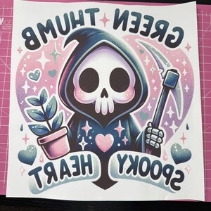 K&ouml;nnte beinhalten: Eine Cartoon-Illustration des Sensenmanns mit einem rosa Herzen, einer Pflanze in einem rosa Topf und dem Text "Green Thumb", "Spooky" und "Heart" in einem rosa und violetten Farbschema.
