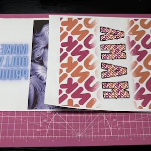 K&ouml;nnte beinhalten: Ein wei&szlig;es Blatt Papier mit dem Text "PROUD DOLLY MAKE" in blauen Neonbuchstaben. Das Papier ist teilweise von einem anderen Blatt Papier mit einem rosa und orangefarbenen Muster und dem Wort "MAMMA" in schwarzen Buchstaben bedeckt.