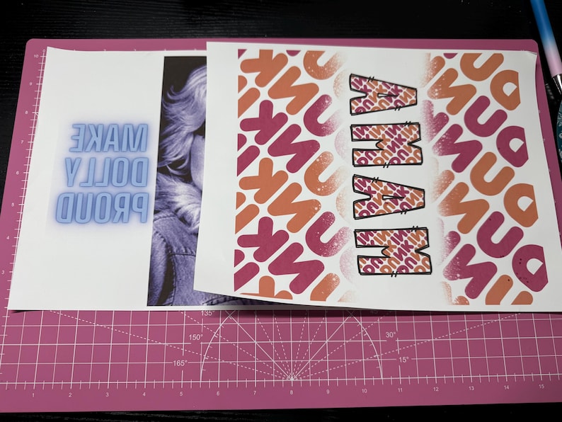 K&ouml;nnte beinhalten: Ein wei&szlig;es Blatt Papier mit dem Text "PROUD DOLLY MAKE" in blauen Neonbuchstaben. Das Papier ist teilweise von einem anderen Blatt Papier mit einem rosa und orangefarbenen Muster und dem Wort "MAMMA" in schwarzen Buchstaben bedeckt.