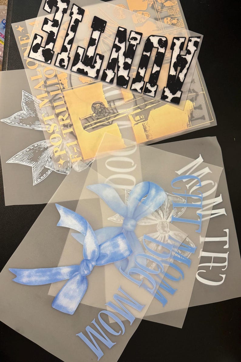 Puede incluir: Tres hojas de papel de transferencia transparente con dise&ntilde;os para planchar. La hoja superior presenta un dise&ntilde;o de estampado de vaca en blanco y negro que deletrea "LOVE". La hoja del medio presenta un dise&ntilde;o amarillo con un patr&oacute;n floral y el texto "Post Malone - 1 Trillion". La hoja inferior presenta un dise&ntilde;o azul con un lazo y el texto "Chill With The Dog Mom".
