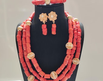 May include: A coral-colored jewelry set displayed on a black mannequin. The set includes a multi-strand necklace, a bracelet, and matching earrings. The necklace and bracelet feature gold-colored accents. The earrings have a floral design.