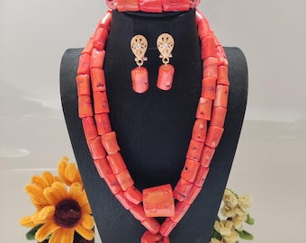 May include: A coral-colored jewelry set displayed on a black mannequin. The set includes a necklace, bracelet, and earrings. The necklace features multiple strands of rectangular beads, with a large square and rectangular pendant. The earrings have gold-colored accents.