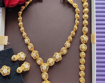 May include: A gold-tone jewelry set with a necklace, bracelet, and earrings. The necklace and bracelet feature a repeating pattern of round, flower-shaped designs with clear stones. The earrings are small and feature a similar flower design.