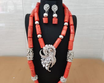 May include: A coral-colored jewelry set displayed on a black stand. The set includes a necklace, bracelet, and earrings. The necklace features large coral beads with silver accents and a decorative pendant. The earrings and bracelet also have silver details.