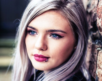 May include: A woman with long, silver-grey hair, wearing dark clothing, looks to the side with a serious expression. She has blue eyes and dark eyeliner. Her lips are painted a deep pink color.
