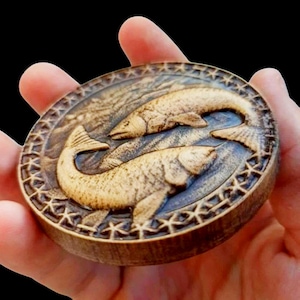 Puede incluir: Una talla ovalada de madera con dos peces en una disposición de yin-yang. Los peces son de color marrón claro, con escamas y aletas detalladas, sobre un fondo azul grisáceo. El borde tiene un patrón geométrico decorativo.