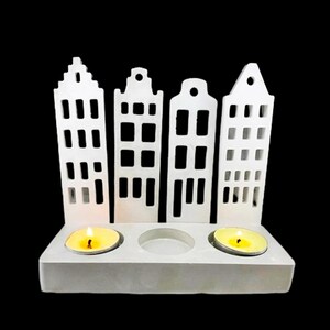 Puede incluir: Portavelas de cerámica blanca con forma de tres casas con tres pozos para velas. El portavelas está sobre un fondo negro.