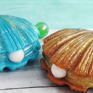 May include: Two decorative seashell-shaped boxes with a pearl inside. One box is blue with gold accents and the other is gold with silver accents.
