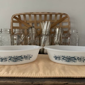 May include: Two white oval baking dishes with blue floral designs. The dishes are on a beige placemat.