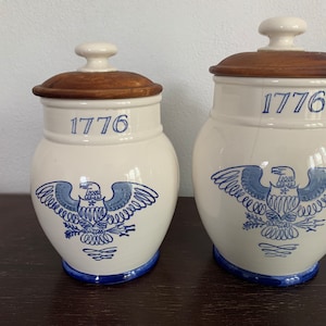 May include: Two white ceramic jars with wooden lids. Both jars feature a blue and white illustration of a bald eagle with the year 1776 printed on the front.
